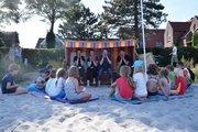 Menschen mit gefalteten Händen in einem Strandkorb, davor sitzt eine Gruppe Kinder im Sand