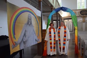 Zwei Frauen in weißer geistlicher Kleidung mit regenbogenbunten Schal stehen unter einem regenbogenbunten Bogen