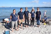 Fünf Menschen in gleichen Shirts schauen zum Betrachter, dahinter zwei Kinder, die im Sand buddeln