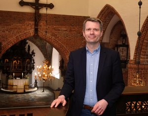 Ein Mann in Jackett und Hemd steht auf der Empore einer Kirche. Im Hintergrund Kreuz und Altar