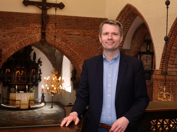 Ein Mann in Jackett und Hemd steht auf der Empore einer Kirche. Im Hintergrund Kreuz und Altar