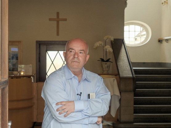 Herr Zander steht in einem hellen Hemd in einem Treppenhaus, die Arme vor der Brust verschränkt, im Hintergrund ein Kreuz an der Wand.
