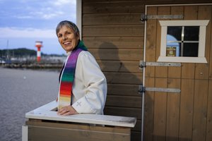 Eine Pastorin steht vor einer kleinen Holzkirche. 