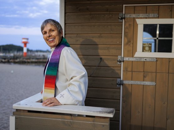 Eine Pastorin steht vor einer kleinen Holzkirche. 