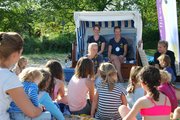 Eine Gruppe Kinder sitzt vor einem Strandkorb, in dem zwei Menschen sitzen