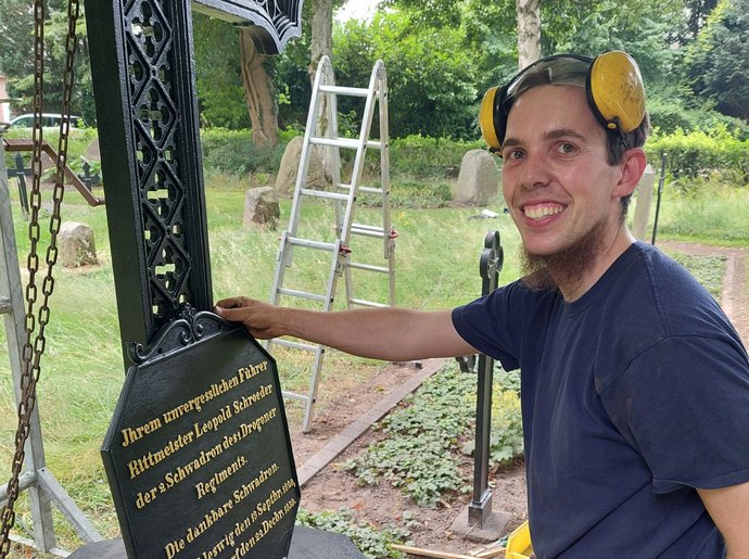 Junger Mann in Arbeitskleidung lächelt in die Kamera, hält schwarze Platte mit goldener Schrift