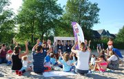 Eine Gruppe Menschen sitzt im Sand und macht nach, was zwei Menschen im Strandkorb vormachen