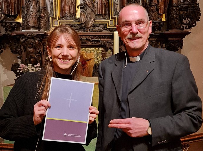 Ein Mann und eine jüngere Frau stehen zusammen vor einem Altar. Die Frau hat eine Urkunde in der Hand.