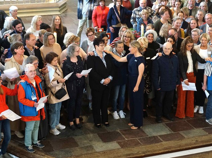Eine Gruppe von Menschen steht im Altarraum einer Kirche. In der Mitte wird eine Frau gesegnet. Die Menschen legen jeweils eine Hand auf die Schulter ihrer Nachbarn.