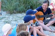Sieben Kinder sitzen im Sand, daneben eine Frau mit Gitarre