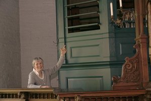 Eine Frau steht neben einer Orgel und zeigt auf Holzlamellen, die halb geöffnet sind.