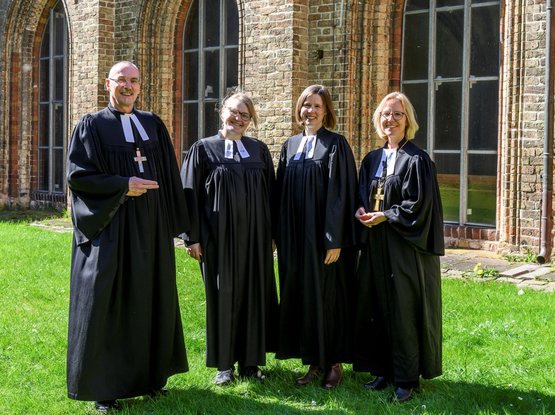 Ein Mann und drei Frauen in geistlicher Kleidung stehen im Sonnenschein auf einer Wiese, im Hintergrund eine Kirche.
