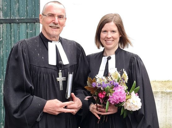 Ein Mann und eine Frau in geistlicher Kleidung (Talar) stehen nebeneinander. Die Frau hält einen Blumenstrauß.