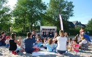 Eine Gruppe Menschen sitzt im Sand und macht nach, was zwei Menschen im Strandkorb vormachen