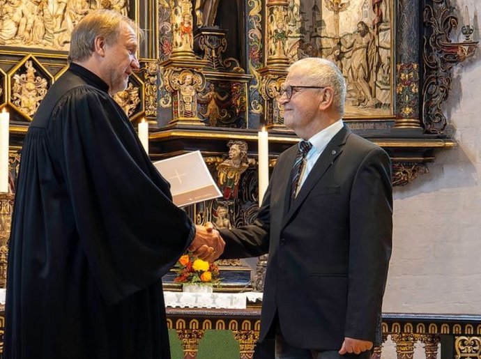 Ein Mann ist geistlicher Kleidung überreicht einem älteren Mann im Anzug eine Urkunde. Im Hintergrund ist ein Altar zu sehen.