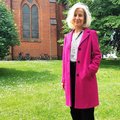 Eine Frau im pinken Mantel steht auf einer Wiese, im Hintergrund ein Kirchgebäude